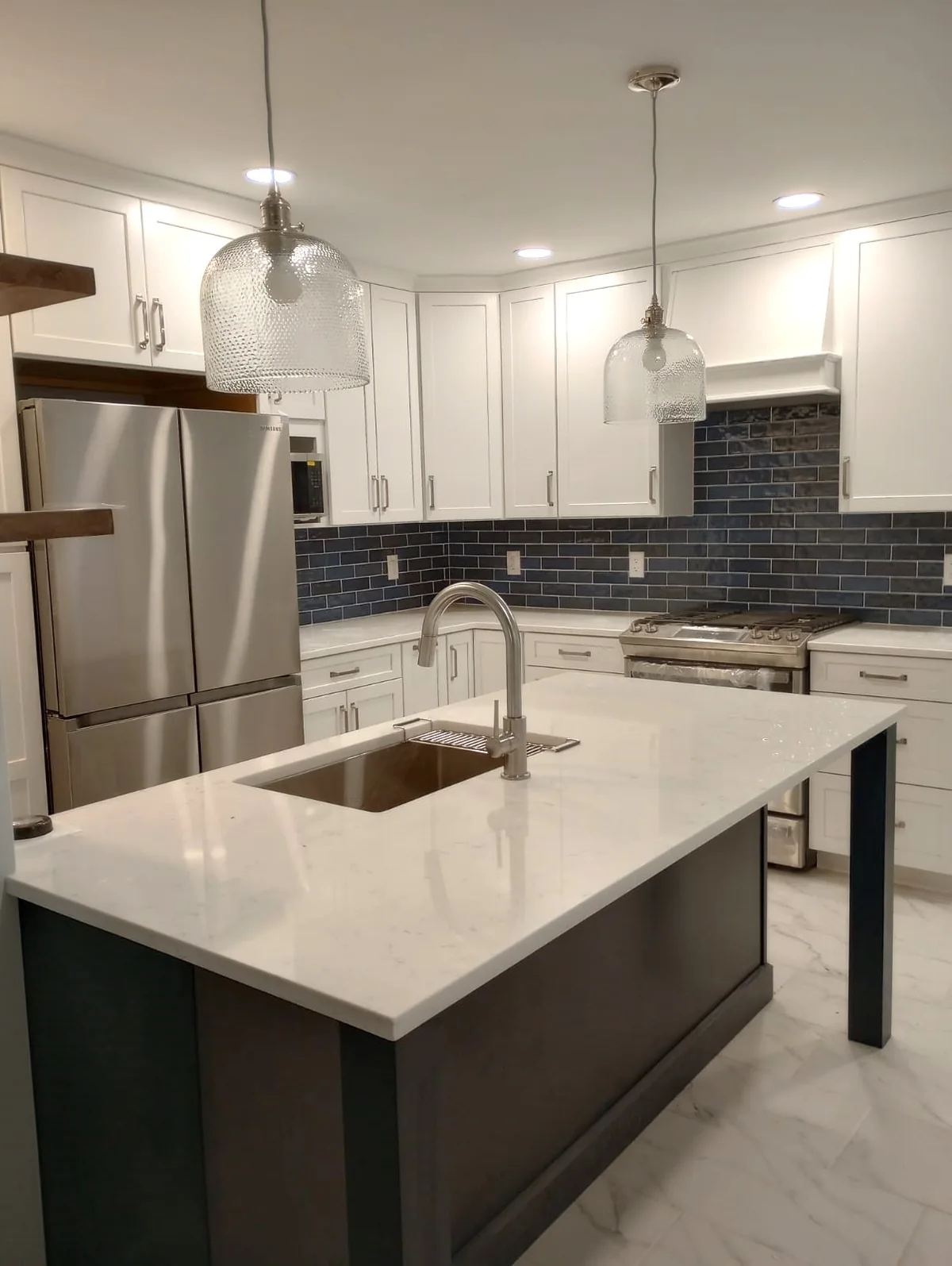 Glass pendant lights above a kitchen island with recessed lights, white cabinets and a dark tile backsplash.